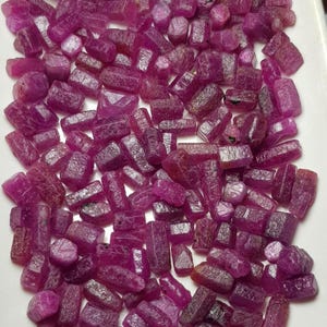 May include: A close-up of a collection of raw ruby gemstones. The stones are a deep pinkish-red hue, with a natural, unpolished appearance. The gemstones vary in size and shape, with a rough, textured surface.