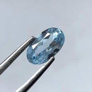 May include: An oval-shaped, light blue gemstone held by silver tweezers. The gemstone has multiple facets that reflect light, creating a sparkling effect. The gem is a pale, sky blue colour.