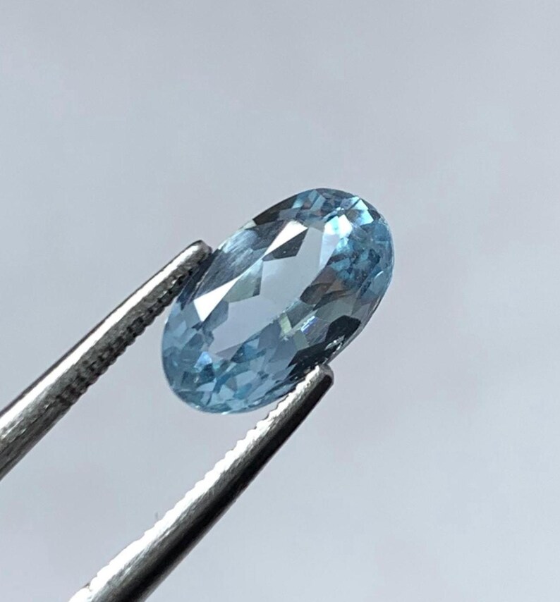 May include: An oval-shaped, light blue gemstone held by silver tweezers. The gemstone has multiple facets that reflect light, creating a sparkling effect. The gem is a pale, sky blue colour.