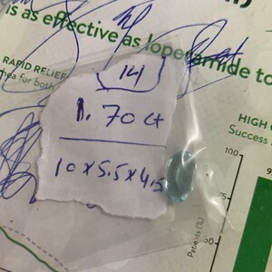 May include: A small, pale blue gemstone in a clear plastic bag. The bag rests on paper with handwritten notes, including measurements of 10x5.5x4.5 mm and a weight of 1.70 ct. The background includes medical information.