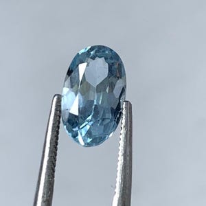 May include: A light blue, oval-cut gemstone is held by tweezers. The gemstone's facets catch the light, displaying its clarity. The background is a soft grey. The gemstone is likely to be measured in millimeters.