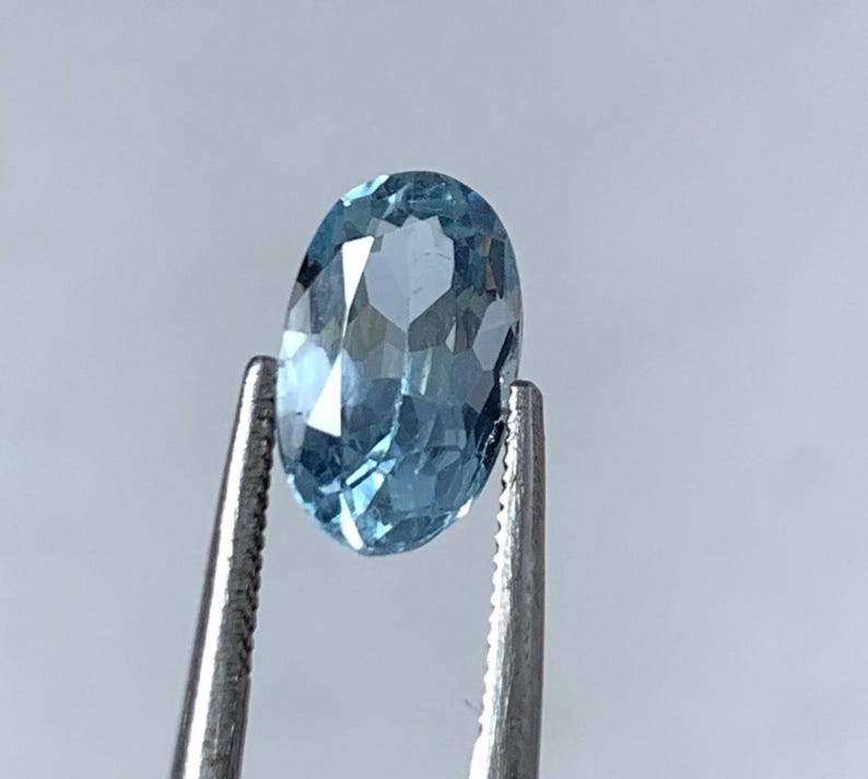 May include: A light blue, oval-cut gemstone is held by tweezers. The gemstone's facets catch the light, displaying its clarity. The background is a soft grey. The gemstone is likely to be measured in millimeters.