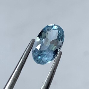 May include: A light blue, oval-cut gemstone is held by silver tweezers. The faceted surface of the gem reflects light, creating a sparkling effect. The background is a soft, neutral grey.