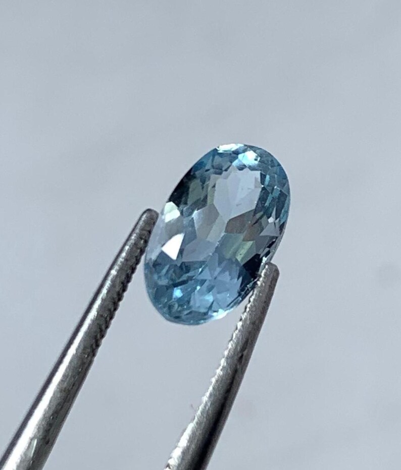 May include: A light blue, oval-cut gemstone is held by silver tweezers. The faceted surface of the gem reflects light, creating a sparkling effect. The background is a soft, neutral grey.