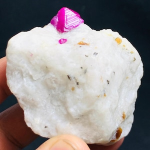 Op de afbeelding: Een ruw, wit kwartsspecimen met een levendige, fuchsia-kleurige robijnkristalformatie. De robijn heeft een kristallijne structuur en zit op het kwarts. Kleine vlekjes van andere mineralen zijn zichtbaar.