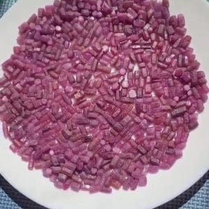 May include: A collection of small, raw, cylindrical ruby gemstones in various shades of deep pink and magenta, scattered across the surface of a plain white circular plate. The stones have a rough, unpolished texture and irregular shapes.