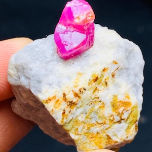 Puede incluir: Un vibrante cristal de rubí rosa se encuentra sobre una formación rocosa blanca y marrón. El rubí es de un rosa intenso con un aspecto facetado. La roca tiene una superficie rugosa y texturizada con áreas amarillas y marrones.