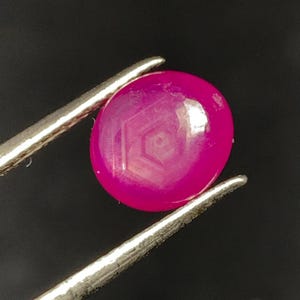 Pode incluir: Uma gema magenta vibrante e redonda é segurada por pinças prateadas contra um fundo preto. A gema polida revela um padrão geométrico sutil. As pinças são posicionadas para exibir a gema.