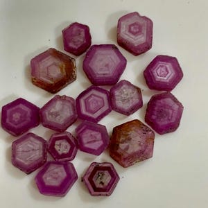 May include: A collection of raw, hexagonal-shaped ruby gemstones. The gems display a deep magenta color with lighter interior patterns. Some stones have a brownish hue on their edges, adding to their natural appearance. The gemstones are scattered across a white surface.