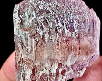 May include: A translucent, crystalline mineral specimen with a pale pink hue. The rough-hewn surface displays a complex, textured pattern with vertical striations. The mineral is held in a person's hand against a black background.