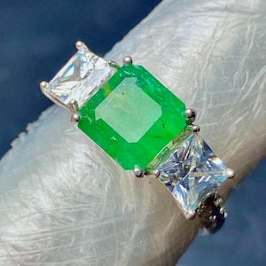 Peut inclure: Une bague en argent avec une grande pierre précieuse vert émeraude entourée de deux petites pierres précieuses claires de forme carrée.