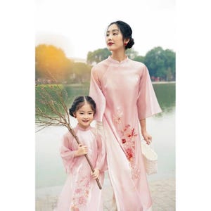 May include: A mother and child are seen wearing matching pink dresses with floral embroidery. The dresses have a mandarin collar and long sleeves. The child is holding a branch with small twigs. The mother is holding a white basket.