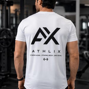 Op de afbeelding: Een wit T-shirt met korte mouwen en een groot zwart AX-logo op de achterkant. Onder het logo staat de tekst ATHLIX, gevolgd door de zin STRONG MIND. STRONG BODY. ONE GOAL. en een klein grafisch icoon van een halter onderaan.