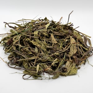 May include: A pile of dried herbs and leaves, featuring various shades of green and brown. The composition includes long, thin stems and small, dried flower heads. The herbs are arranged on a white surface, creating a natural, earthy aesthetic.