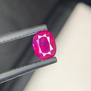 May include: A vibrant, oval-cut ruby gemstone held by tweezers. The ruby displays a deep, saturated pink-red hue with internal facets creating a starburst pattern. The gem is set against a neutral background.