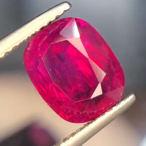 Pode incluir: Uma gema de rubi facetada, em forma oval, segurada por uma pinça. O rubi é de uma cor vermelha profunda e vibrante, com múltiplas facetas que refletem a luz. A pinça é prateada.