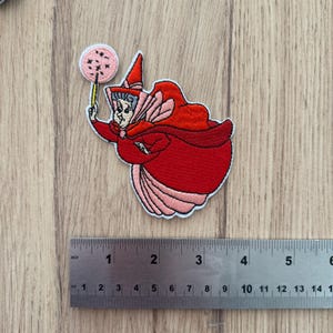Puede incluir: Parche bordado con un personaje de dibujos animados con capa y sombrero rojos, sosteniendo una varita con un círculo rosa. El parche mide aproximadamente 7,6 cm de largo.