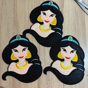 May include: Three felt appliques of a cartoon princess with black hair, a blue headband, and yellow earrings and necklace. The princess has a fair complexion and a red smile. The appliques are arranged on a light brown wooden surface.