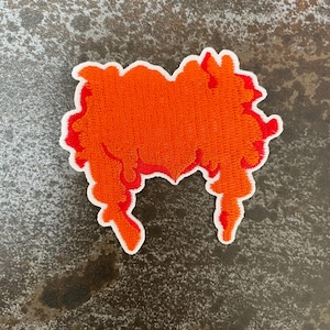 Pode incluir: Remendo bordado com um penteado estilizado laranja e vermelho com uma borda branca. O remendo tem uma aparência texturizada, com o cabelo laranja ocupando a maior parte do design. O remendo é provavelmente para roupas ou acessórios.