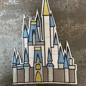 Könnte beinhalten: Gestickter Aufnäher mit einem stilisierten Schloss mit mehreren Türmen und Türmchen. Das Schloss hat blaue, goldene, graue und weiße Details mit einer blauen Flagge an der Spitze. Der Aufnäher hat einen weißen Rand.