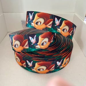 Peut inclure: Un rouleau de ruban vert, marron et blanc avec un motif répété d'un cerf de dessin animé avec un papillon. Le cerf a une fourrure brune et de grands yeux bruns.