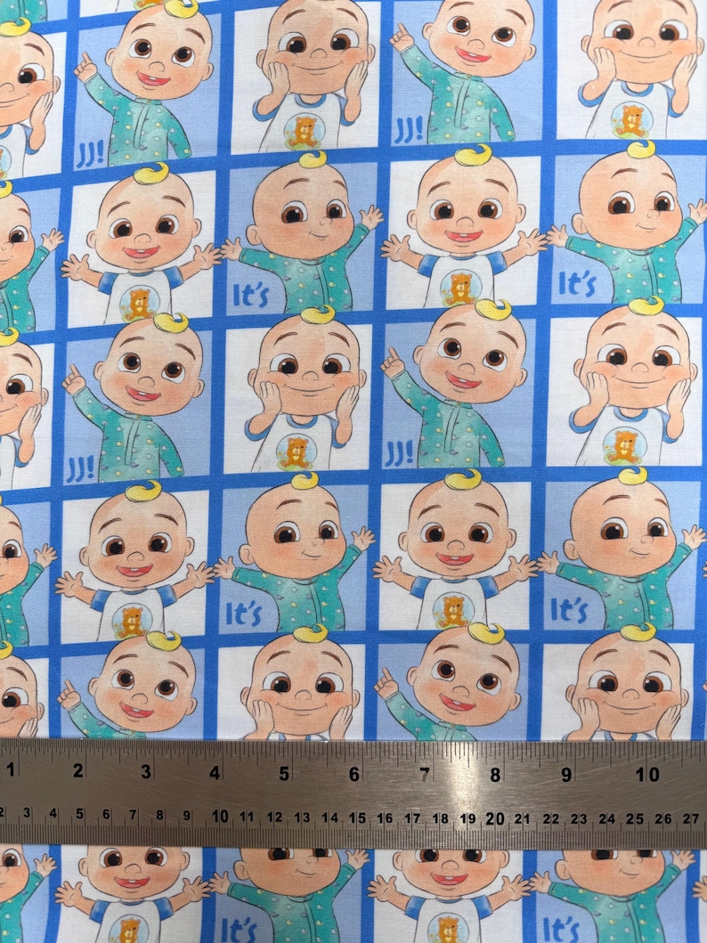 May include: A blue and white patterned fabric featuring a repeating grid of cartoon characters in green pajamas, with some squares displaying the text "It's". A metal ruler is positioned at the bottom, showing measurements up to 10.5 inches.
