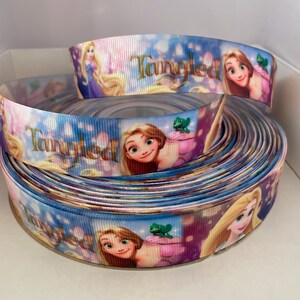 Puede incluir: Cinta enrollada con la palabra "Tangled" e imágenes de Rapunzel de la película de Disney. La cinta tiene un fondo azul, morado y rosa con la cara de Rapunzel y otros personajes. La cinta está enrollada en una caja blanca.