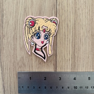 Könnte beinhalten: Gestickter Aufnäher mit dem Gesicht einer Zeichentrickfigur mit blonden Haaren, blauen Augen und einem rosa Hintergrund. Der Aufnäher ist etwa 5 cm breit und 5 cm hoch. Der Aufnäher befindet sich auf einer Holzoberfläche.