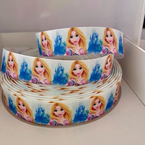 May include: White satin ribbon with a repeating pattern of a cartoon image of Rapunzel with long blonde hair, a pink dress, and a blue background.