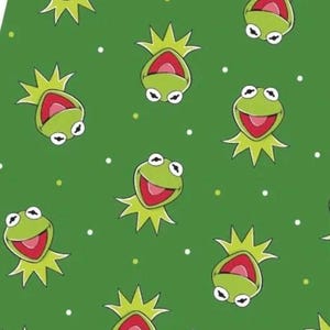 May include: A green fabric pattern featuring a repeating graphic of a smiling frog character with a pointed collar. The design includes small white and light green dots scattered across the background, creating a consistent and uniform visual texture.