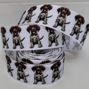 May include: A roll of white grosgrain ribbon featuring a repeating pattern of brown and white spotted puppy illustrations. The puppies are depicted in a sitting position with their tongues visible. The ribbon has a textured, ribbed surface.