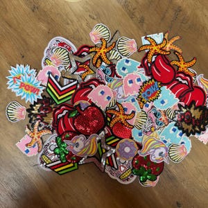 Può includere: Una collezione di toppe colorate e con paillettes con vari disegni, tra cui una mela rossa, stelle marine, labbra, un unicorno e un teschio. Alcune toppe presentano la parola "POW!" in stile fumetto. Le toppe sono su una superficie di legno.