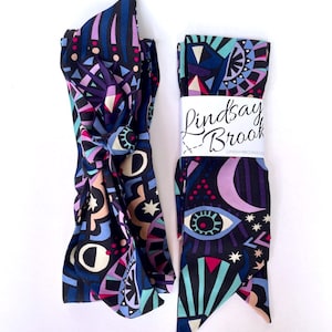 May include: Two black fabric headbands with a colorful geometric pattern featuring eyes, stars, and moons. The headband on the right has a white label with the text "Lindsay Brook" and "Lindsaybrookdesign.co".