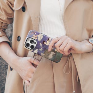 May include: A phone case with a whimsical design featuring a purple and blue background with illustrations of figures, clouds, and flowers. The case has a gold-colored border and a camera cutout.