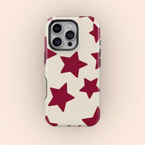 Puede incluir: Funda de teléfono con fondo crema y un patrón de estrellas rojo oscuro. La funda tiene un acabado brillante y un módulo de cámara negro. Las estrellas varían en tamaño y están distribuidas uniformemente.