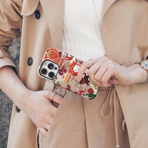 May include: A patterned phone case featuring a strawberry, a bear, and a butterfly design. The case has a patchwork design with red, white, and brown colors. The phone case is held by a person wearing a tan trench coat.