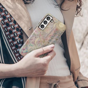 May include: A floral patterned phone case in shades of pink, yellow, and green. The case features a sun and moon design and is held in a person's hand. The phone case is designed for a smartphone with three camera lenses.