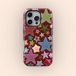 以下が含まれることがあります： さまざまな色の星の形が散りばめられた、バーガンディ色の背景の携帯電話ケース。星はピンク、青、緑、金の色合いで、キラキラした質感のものもあります。ケースには黒い縁とカメラの切り抜きがあります。