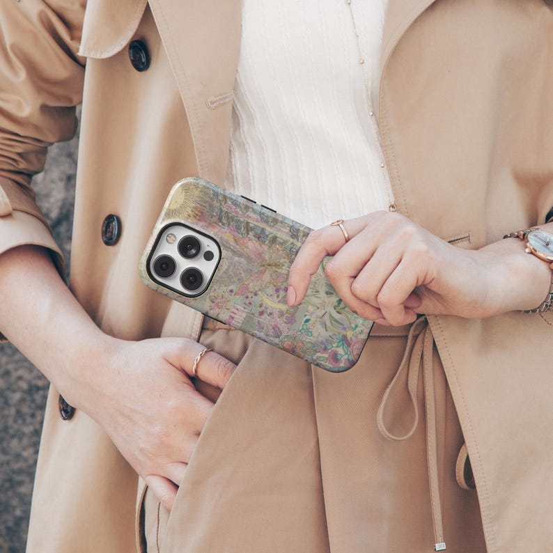May include: A smartphone case with a colorful floral design in shades of pink, green, and yellow. The case is held in a person's hand, with a tan trench coat in the background.