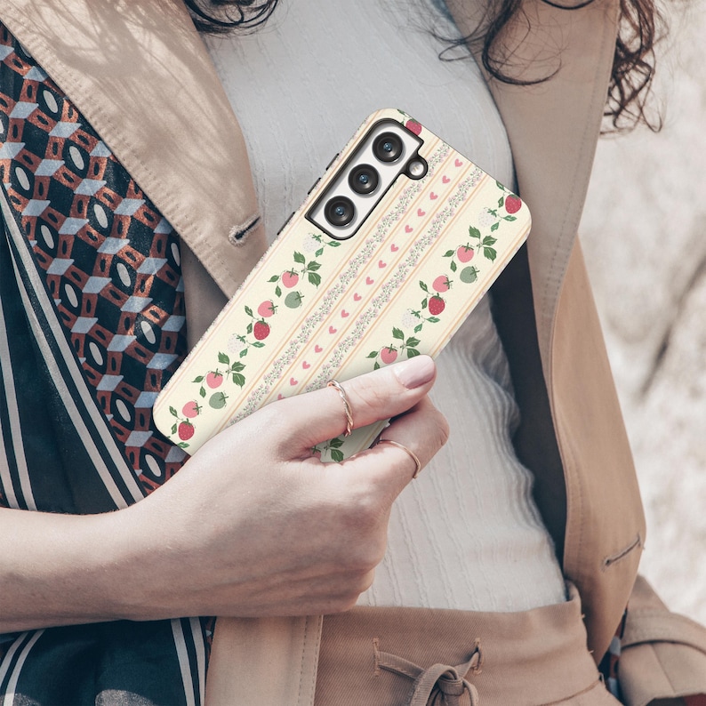 Peut inclure: Une coque de t&eacute;l&eacute;phone blanche avec un motif floral de fraises roses et vertes et de c&oelig;urs. La coque a un d&eacute;coupage pour l'appareil photo et un fond blanc.