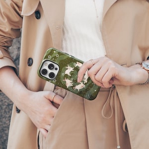 Puede incluir: Una funda de tel&eacute;fono verde con un dise&ntilde;o de conejos blancos y estrellas. La funda tiene un fondo verde oscuro con un dise&ntilde;o en espiral. La persona lleva una gabardina beige y una camiseta blanca.