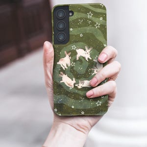 Puede incluir: Funda de tel&eacute;fono verde con un dise&ntilde;o de conejos blancos en espiral, con estrellas blancas. La funda tiene un acabado mate y un recorte para una c&aacute;mara de triple lente.