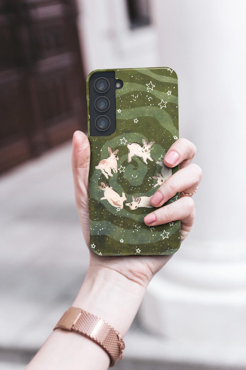Puede incluir: Funda de tel&eacute;fono verde con un dise&ntilde;o de conejos blancos en espiral, con estrellas blancas. La funda tiene un acabado mate y un recorte para una c&aacute;mara de triple lente.