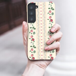 Peut inclure: Une coque de t&eacute;l&eacute;phone blanche avec un motif de tomates roses et rouges, de feuilles vertes et de fleurs blanches. La coque a un motif de rayures verticales avec de petits c&oelig;urs.