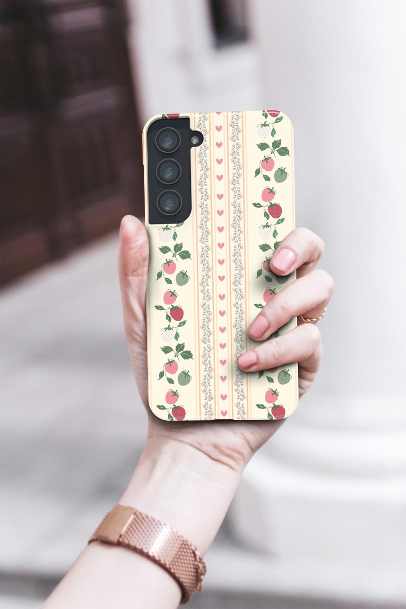 Peut inclure: Une coque de t&eacute;l&eacute;phone blanche avec un motif de tomates roses et rouges, de feuilles vertes et de fleurs blanches. La coque a un motif de rayures verticales avec de petits c&oelig;urs.