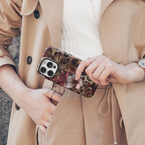 May include: A smartphone case featuring a floral design with burgundy, pink, and white flowers, set against a leopard print background. The case is held by a person wearing a tan trench coat.