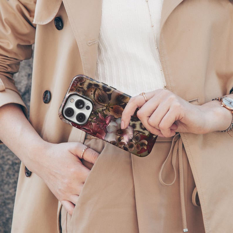 May include: A smartphone case featuring a floral design with burgundy, pink, and white flowers, set against a leopard print background. The case is held by a person wearing a tan trench coat.