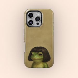 Puede incluir: Una funda de teléfono de color canela con una rana verde con un corte de pelo bob marrón oscuro. La funda del teléfono tiene una cámara de triple lente y un flash. La rana tiene ojos grandes y redondos y una expresión neutra.