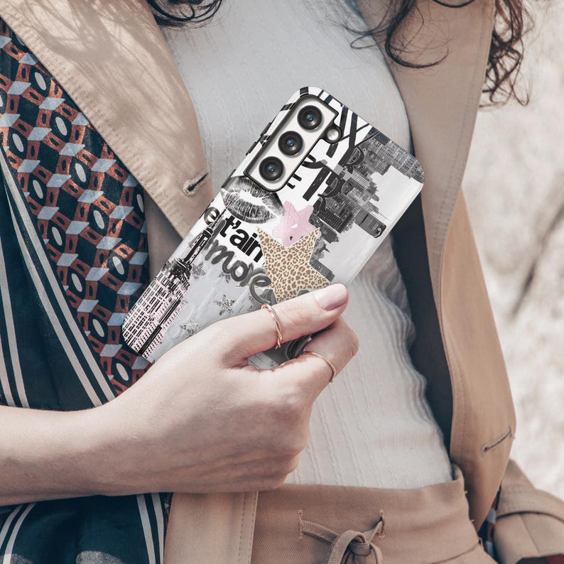 May include: A white phone case with a black and white collage design featuring the Eiffel Tower, lips, and text. Accents include a pink star and a leopard print shape. The case is held by a person.