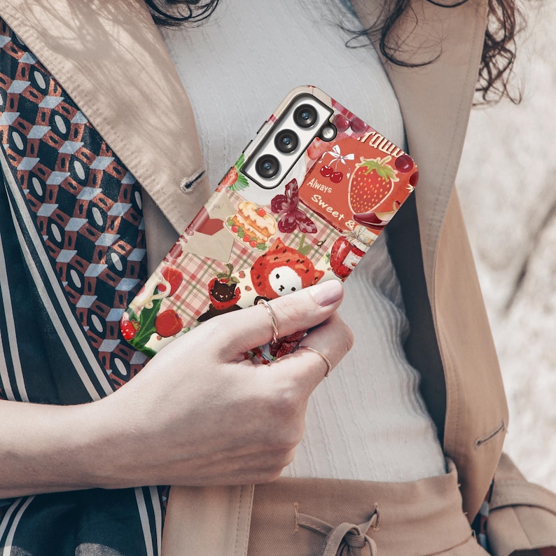 May include: A colorful phone case featuring a patchwork design with strawberries, cherries, and other fruit illustrations. The case has the words "Always Sweet" and "raww" printed on it. The case is held by a person.